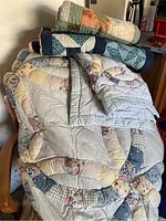 Photo showing a stack of quilts and pillow shams on a wooden chair. Quilts have floral and patchwork patterns with various pastel colors.