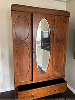 Front view of wooden wardrobe cabinet with three paneled doors and oval mirror on center door. Bottom drawer is open, showing storage space.