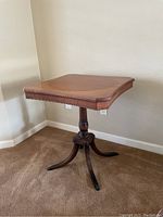 Photo of a wooden side table with a single pedestal and three curved legs, showing the top and base in a corner against carpeted floor and wall.