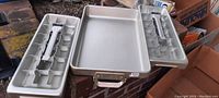 Set of three antique ice cube trays made of aluminum displayed on a brick surface. Two are traditional trays with lever mechanism and compartments, and one is a simple rectangular tray.