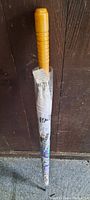 Full view of the closed umbrella with a wooden handle, wrapped in plastic and sealed showing the comic strip print.
