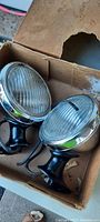 Two headlights in original cardboard box seen from an angled top view showing fluted glass lenses and steel mounts
