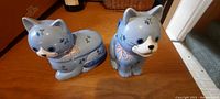 Two blue glass cat figurines, one lying down and one sitting, both with blinky eyes and painted floral details, shown on wooden table.
