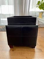 Side view of the black ottoman with faux leather upholstery showing stitching details and wooden legs.