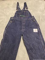 Full view of the dark blue denim bib overalls laid flat showing front bib pocket, straps, and metal buttons.