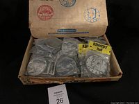 Full view of the original cardboard box containing various round metallic belt buckles sealed individually in plastic packages. Visible belt buckles show Buffalo Nickel and Kennedy Half Dollar designs. The lid of the box images U.S. coin graphics in red and blue.