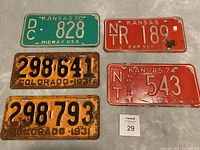 Photo showing all five plates on a surface: 2 orange 1931 Colorado plates with large black numbers, 1 green Kansas 1970 plate, and 2 red Kansas plates, one with expiration SEP 71 and one labeled 'Truck', showing rust and wear.