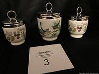 Front view of all three porcelain egg coddlers with silver lids, showing painted bird and blackberry designs.