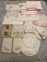 Overview of 15 embroidered small linens and decorative pieces of various shapes, primarily square and oval, laid out on a surface showcasing different floral embroidery patterns and crochet edged borders.