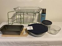 Photo showing Anchor Hocking rectangular 3 QT glass pan set on green metal plate and cup rack, blue Stanley thermos, metal loaf pan, two Pyrex glass bowls (one covered with blue lid), and wooden cutting board beneath.