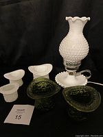Milk glass hobnail lamp with ruffled edge, three milk glass top hat vases, and two green glass top hat vases arranged together on a dark background, showing the textures and shapes of all items.
