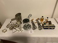 Overview of vintage metal kitchenware and collectibles on white table including flour sifter, egg beaters, cookie cutters, bottles, shaving brushes, and candle holder