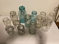 Front and side view of all 13 older glass jars arranged on a white surface showing different sizes, colors (clear and blue), and bale wire handles with rust.