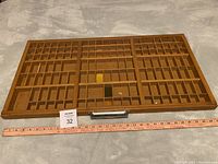 Wooden printers tray laid flat horizontally showing the front side with many small compartment divisions and a metal handle on one edge. Visible scratches and stains on wood surface.