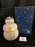 Front view of the ceramic snowman cookie jar next to its blue box with gold stars and product label.