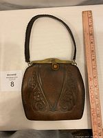 Front view of vintage brown tooled leather handbag, showing floral pattern, braided handle, and metal clasp with decorative accent.