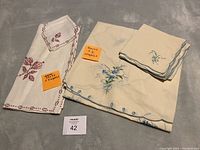 Both sets of tablecloths and napkins laid out flat with sticky notes indicating quantity and type, showing red embroidered square set on the left and blue floral round set on the right.