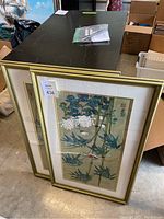 Two framed Asian inspired floral and bamboo art pieces resting against a black table, gold frames with beige mats.