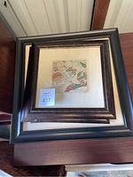 Top view showing three square framed art pieces stacked, dark frames, matted nature-themed artworks with visible frame damage.