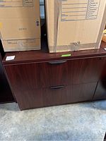 Wooden two-drawer filing cabinet shown with two cardboard boxes on top, view of front and handles.