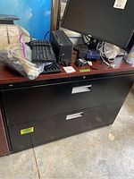 Black metal two-drawer filing cabinet under a wooden desk with office equipment on top.