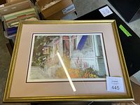 Framed art print showing a colorful porch with flowers and a hanging sign, gold frame and pink mat, displayed on a table with boxes in the background.