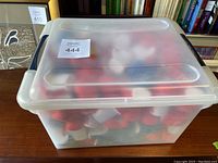 Large translucent plastic storage bin filled with red and blue punching bag shaped stress balls on a wooden surface in front of a bookshelf.