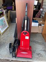Red Simplicity upright vacuum cleaner with handle and hose attachment, placed on concrete floor next to two black accessory boxes and furniture items.
