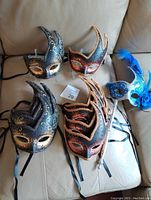 Five handmade and hand-decorated Venetian masks arranged on couch showing colors, horn-like extensions, and ornate decorations
