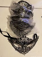 Five large ornate black wire masquerade masks with rhinestone decoration, tied with black satin ribbons, stacked in clear plastic bags.