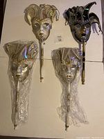Three handheld Venetian jester masks arranged on a white surface, two silver/gold wrapped in plastic, one black/silver/gold with braided trim and handle