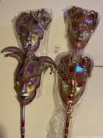 Four handheld jester masquerade masks wrapped in plastic, 2 in purple and 2 in red color.