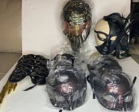 Group photo of masks on table with plastic wrapping visible. Includes long nose masks, black feather mask, handheld masks, and burgundy half moon masks.