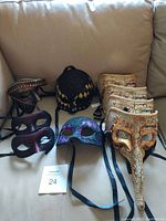 Photo showing a variety of masquerade masks on a beige couch including long nose gold masks, black and purple glitter masks, and bronze metallic masks, each with black ribbons for securing.