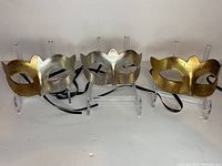 Three silver and gold Venetian style masquerade masks shown on clear display stands with black ribbon ties.