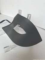 Single black masquerade mask shown from front angle on clear stand highlighting smooth matte resin surface and oval eye hole.