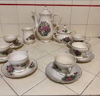 Full set displayed on white tiled surface: coffee pot, creamer, sugar bowl, six cups and six saucers with floral design and gold trim.