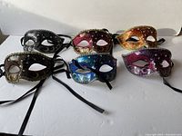 Overview of six Venetian opera-style masquerade masks showing black, red, gold, bronze, blue, and purple masks with glitter, rhinestones, and black tie ribbons.
