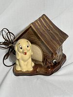 Photo showing ceramic doghouse with attached dog figurine and electrical cord coiled nearby.