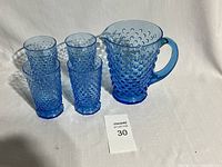 One aqua blue hobnail pattern glass pitcher with handle and four matching tumblers arranged on white surface