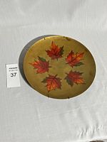 Full view of the circular copper enamel plate on white fabric background showing six maple leaf motif.