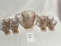 Set of a pink depression glass pitcher and 8 matching glasses with cherry blossom pattern, displayed in natural light showing detailed floral embossing.