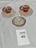 Photo showing two pink Jeanette glass octagonal candle holders and one round pink glass coaster with floral star pattern, all under sunlight on a white surface.