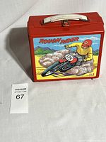 Red plastic vintage lunch box with Rough Rider motocross graphic and white handle shown from front and top angles.