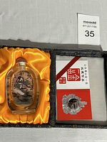 Full view of snuff bottle resting on yellow silk interior of box, with box partially open showing a red and white card and metal ornament.