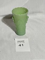 Front and side view of a light green jadeite glass vase featuring a scalloped rim and raised floral designs on the sides, sitting on a fabric surface with a numbered lot card.