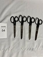 Full view of four vintage steel scissors with black handles, arranged side-by-side on a white textured fabric, showing general used condition and paint chipping on handles.
