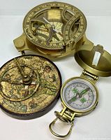Three vintage compasses displayed on a white background. The largest is a brass sundial compass with glass face, Roman numerals, and adjustable measuring scales.