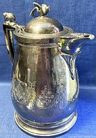 Front angle view of the large antique silver plated coffee pot showing detailed engraved floral garland, animal handle, bird lid finial, and spout design.