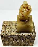 Front view of carved Buddha stamp atop the original patterned box, showing the Buddha figure and detailed box fabric design.